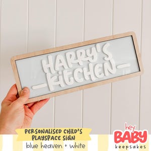 May include: A rectangular wooden sign with the words "HARRY'S KITCHEN" in white, raised lettering. The sign has a light blue background and is framed in natural wood. Text at the bottom reads "PERSONALISED CHILD'S PLAYSPACE SIGN" and "blue heaven + white".