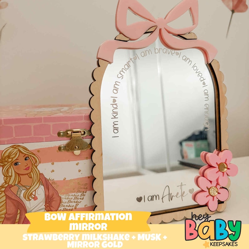 May include: A decorative mirror with a pink bow and flower accents. The mirror has a scalloped edge and includes the text "I am kind, smart, brave, loved, enough" and "I am Arete". The mirror is part of the "Hey Baby Keepsakes" collection.