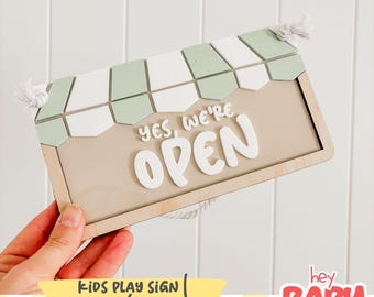 Pode incluir: Uma placa retangular de madeira com fundo bege claro e o texto em relevo branco YES, WE'RE OPEN. A parte superior assemelha-se a um toldo de loja com azulejos alternados em verde sálvia e branco. Uma corda branca está presa aos cantos.