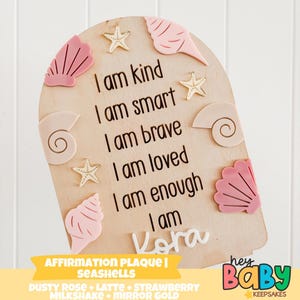 以下が含まれることがあります： 貝殻の装飾が施された木製のアファメーションプラーク。「私は親切で、賢く、勇敢で、愛され、十分です、私は」という言葉と「Kora」という名前が書かれています。貝殻とヒトデが取り付けられています。