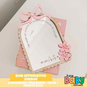 May include: A pink bow-shaped affirmation mirror with a scalloped edge, featuring the phrases "I am kind, smart, brave, loved, enough." Two pink flowers and the text "I am Arete" are on the mirror. The mirror is on a pink box.