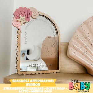 Op de afbeelding: Houten spiegel in de vorm van een zeeschelp met een geschulpte rand. De spiegel heeft de tekst "I am kind, I am smart, I am brave, I am loved, I am enough" en "I am Aria". Versierd met een roze zeeschelp, zeester en een krul.