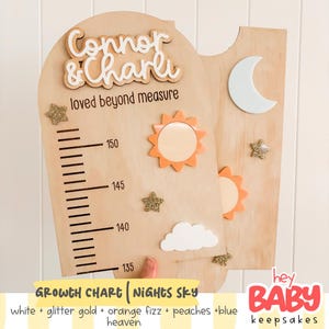 Può includere: Un metro di crescita in legno con i nomi Connor & Charli e il testo loved beyond measure. La tavola include una scala di misurazione da 135 a 150, decorata con una mezzaluna bianca, soli arancioni, stelle glitterate dorate e una nuvola bianca.