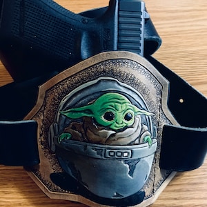 May include: A black handgun rests on a brown leather belt buckle featuring a green and brown image of a character in a pod. The buckle has a gold-coloured border and is set against a light brown wooden surface.