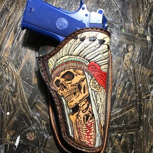 Puede incluir: Una funda de cuero con un diseño de calavera y plumas. La funda es marrón con detalles rojos, blancos y negros. Una pistola azul está dentro de la funda.