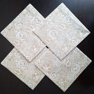 May include: Four square cloth napkins with a floral pattern in beige and white. The napkins are arranged in a diamond shape, with each corner touching. The floral design features detailed flowers and leaves.