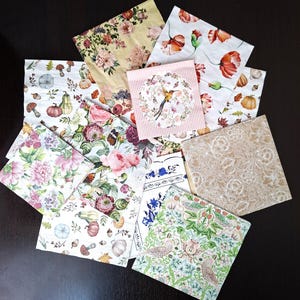 Op de afbeelding: Een verzameling decoratieve papieren servetten met diverse bloemen- en botanische ontwerpen. De servetten tonen afbeeldingen van rozen, klaprozen, pompoenen, vogels en andere natuurlijke elementen in roze, gele en groene tinten. De servetten zijn gerangschikt op een donkere ondergrond.