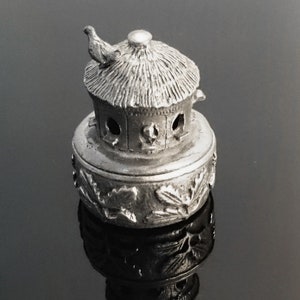 May include: A silver metal birdhouse with a thatched roof and a bird perched on top. The birdhouse is decorated with leaf-like designs around the base.