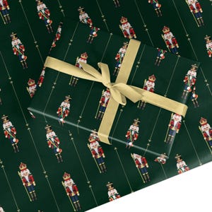 May include: A rectangular gift box wrapped in dark green paper featuring a repeating pattern of nutcracker figures in red, blue, and gold uniforms. The box is tied with a wide, smooth, light gold ribbon arranged in a cross pattern.