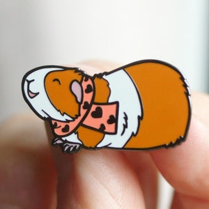 May include: A cartoon enamel pin of a brown and white guinea pig wearing a pink and black heart patterned scarf. The guinea pig is smiling and has its eyes closed.