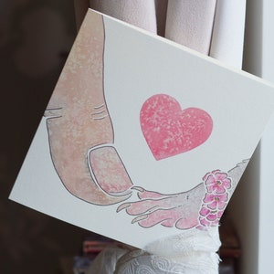 Puede incluir: Un lienzo cuadrado con una ilustraci&oacute;n en acuarela de un pulgar humano grande tocando la mano de una peque&ntilde;a criatura con garras. Un coraz&oacute;n rosa texturizado se sit&uacute;a entre ambas figuras y la criatura lleva una pulsera de flores rosas.