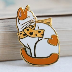 May include: A gold-colored cat enamel pin with a white body, orange accents, and a scarf patterned with black hearts. The pin has a gold border and is set against a blurred background.
