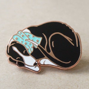 May include: A black and white enamel pin of a sleeping dog wearing a blue and white star patterned bandana. The pin is made of rose gold metal.