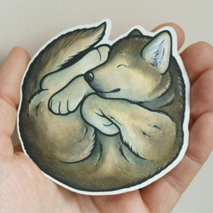 May include: A brown and gray wolf pup curled up sleeping. The pup has a white patch on its chest and a white nose. The sticker is on a white background.