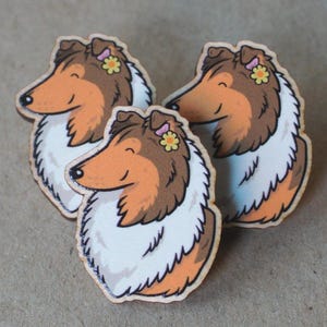 May include: Three wooden pins featuring a stylized illustration of a brown and white collie dog. Each dog has a small yellow flower tucked behind its ear. The pins are shaped to follow the outline of the dog's head and neck, showing a side profile.
