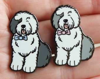 May include: Two enamel pins of Old English Sheepdogs wearing bow ties. The dog on the left is wearing a blue bow tie and the dog on the right is wearing a pink bow tie.