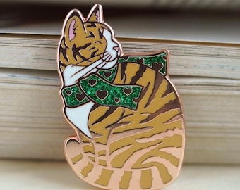 Puede incluir: Un pin de esmalte de oro rosa de un gato atigrado con una bufanda verde de purpurina con corazones. El gato está enroscado en una posición para dormir.
