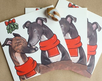 Puede incluir: Un conjunto de tarjetas de felicitación blancas con una ilustración de dibujos animados de dos galgos con bufandas rojas brillantes. Sobre los perros, se representa una rama de muérdago verde con un lazo rojo. Las tarjetas están apiladas.