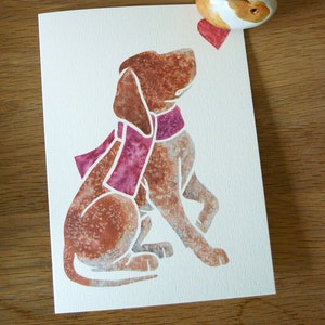 May include: A watercolor illustration of a brown and white dog wearing a pink scarf. The dog is sitting with its head tilted to the side. A small ceramic figurine of a rabbit holding a red heart is in the top right corner of the image.