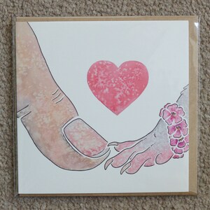Puede incluir: Una tarjeta de felicitaci&oacute;n cuadrada con una ilustraci&oacute;n en acuarela de un pulgar humano tocando la mano con garras de una criatura. Un coraz&oacute;n rosa est&aacute; centrado sobre las manos y la criatura lleva una pulsera de flores rosas en la mu&ntilde;eca.