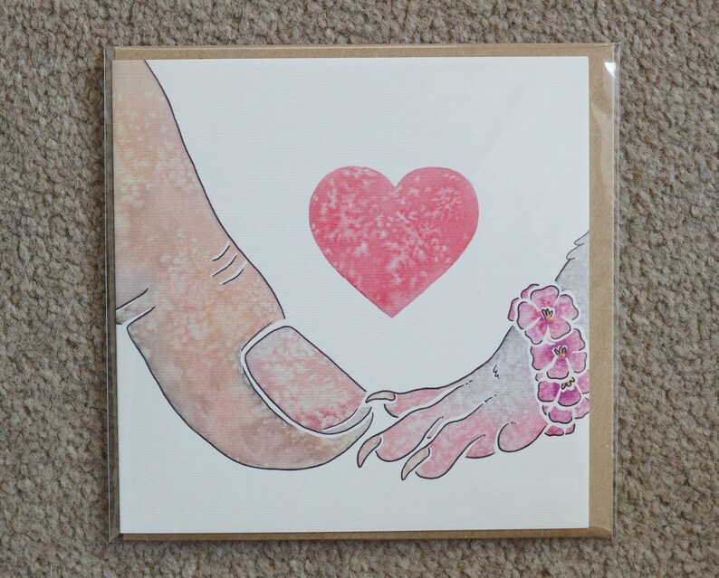 Puede incluir: Una tarjeta de felicitaci&oacute;n cuadrada con una ilustraci&oacute;n en acuarela de un pulgar humano tocando la mano con garras de una criatura. Un coraz&oacute;n rosa est&aacute; centrado sobre las manos y la criatura lleva una pulsera de flores rosas en la mu&ntilde;eca.
