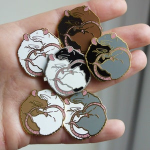 Six different colour variations of a hugging rat enamel pin, all resting on a hand.