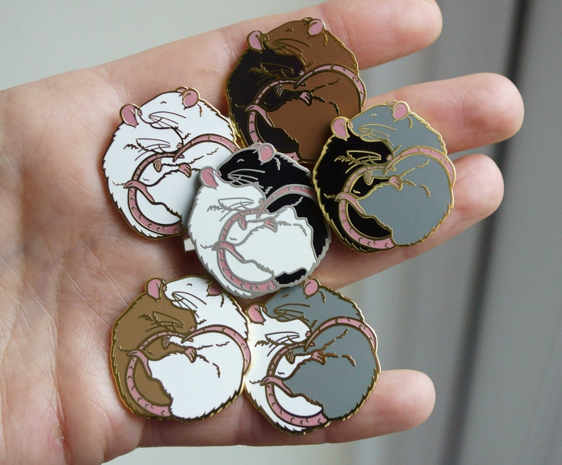 Six different colour variations of a hugging rat enamel pin, all resting on a hand.