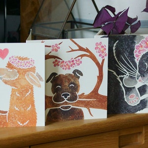 Puede incluir: Tres tarjetas de felicitaci&oacute;n cuadradas con ilustraciones en acuarela. La primera muestra dos alpacas marrones con coronas de flores rosas y corazones. La segunda representa un perro marr&oacute;n sosteniendo una rama con flores rosas. La tercera muestra un gato negro.