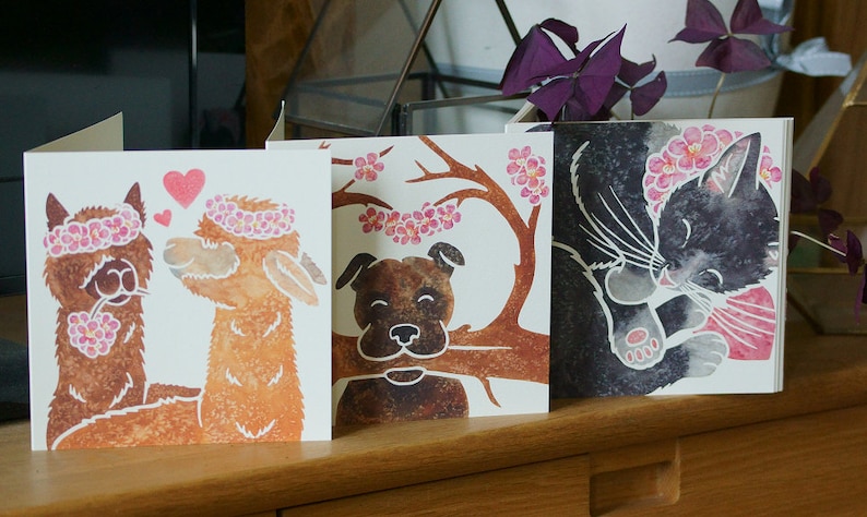 Puede incluir: Tres tarjetas de felicitaci&oacute;n cuadradas con ilustraciones en acuarela. La primera muestra dos alpacas marrones con coronas de flores rosas y corazones. La segunda representa un perro marr&oacute;n sosteniendo una rama con flores rosas. La tercera muestra un gato negro.