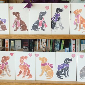 May include: A set of 10 greeting cards featuring different dog breeds wearing scarves. Each card has a pink heart in the top right corner. The cards are displayed on a shelf with books behind them.