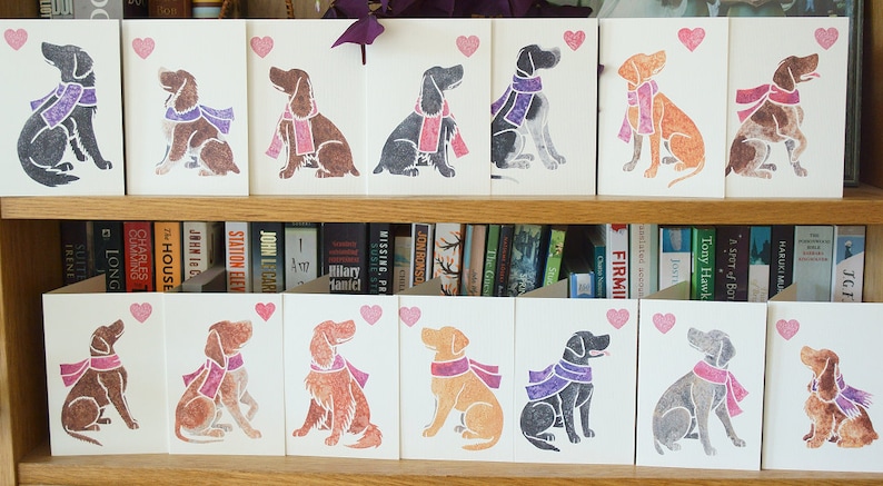 May include: A set of 10 greeting cards featuring different dog breeds wearing scarves. Each card has a pink heart in the top right corner. The cards are displayed on a shelf with books behind them.