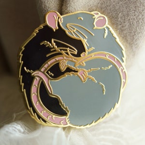 A circular enamel lapel pin of two rats hugging. The left hand side rat is black and the right hand side is blue/grey. The metal plating colour is gold.