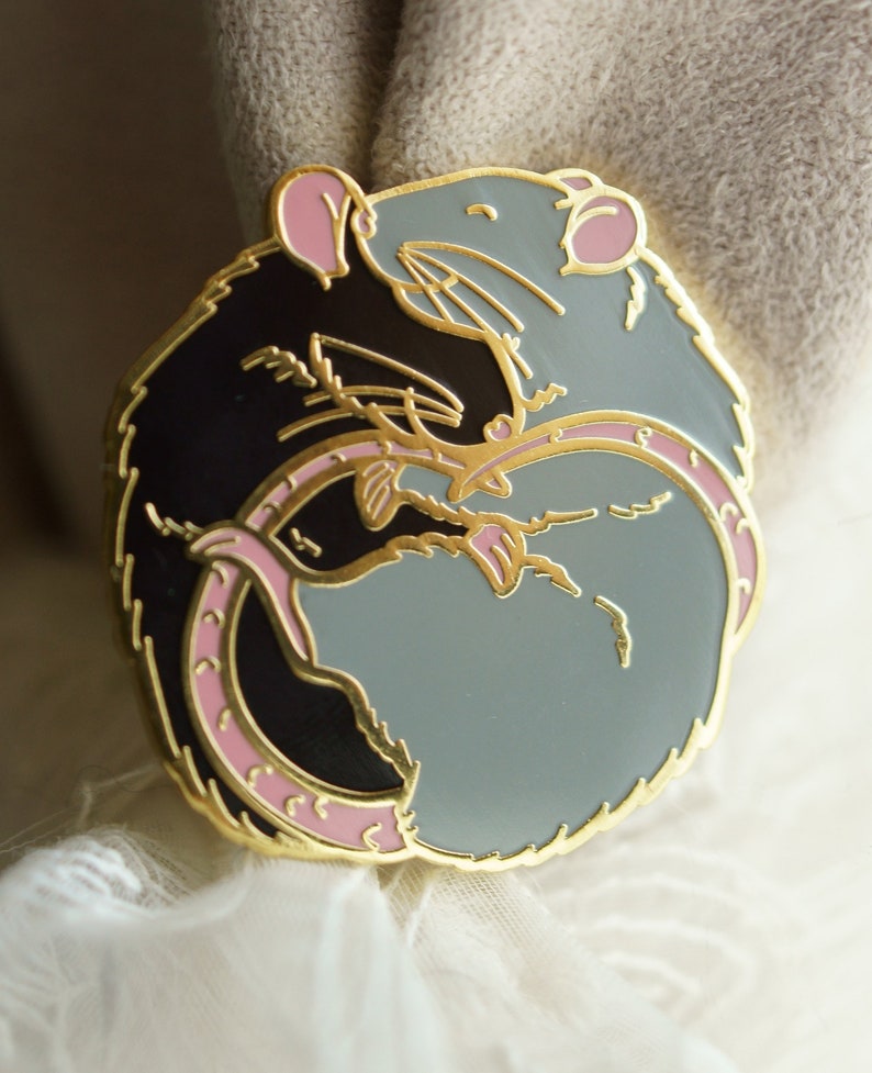 A circular enamel lapel pin of two rats hugging. The left hand side rat is black and the right hand side is blue/grey. The metal plating colour is gold.