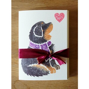 May include: A greeting card with a black and brown dog wearing a purple scarf. The dog is looking up at a pink heart. The card is tied with a burgundy ribbon.