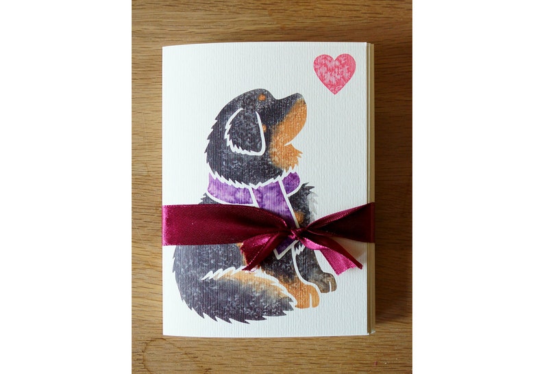 May include: A greeting card with a black and brown dog wearing a purple scarf. The dog is looking up at a pink heart. The card is tied with a burgundy ribbon.