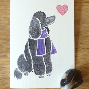 May include: A black poodle with a purple scarf looking at a pink heart. The image is on a white card.