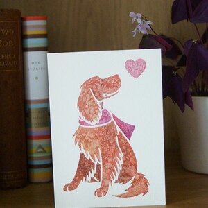 May include: A white card with a pink heart and a watercolor illustration of a brown dog looking up at the heart. The dog is wearing a pink scarf.