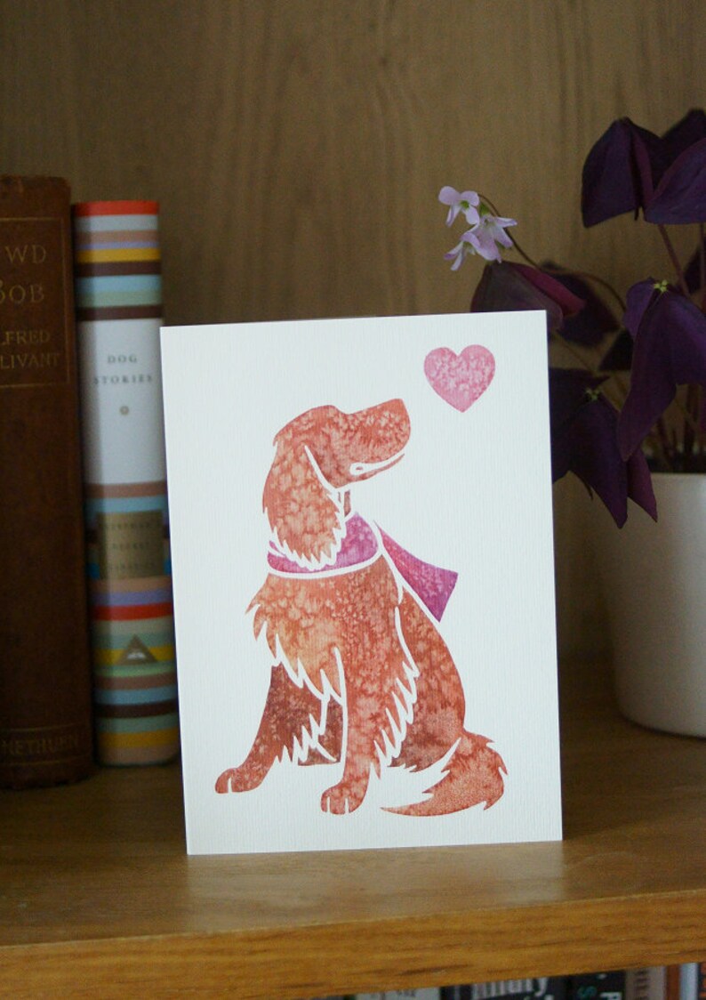 May include: A white card with a pink heart and a watercolor illustration of a brown dog looking up at the heart. The dog is wearing a pink scarf.