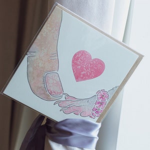 Puede incluir: Una tarjeta de felicitaci&oacute;n cuadrada con una ilustraci&oacute;n en acuarela de un pulgar grande tocando una mano m&aacute;s peque&ntilde;a con garras. Un coraz&oacute;n rosa texturizado se sit&uacute;a entre ambas manos, y la mano peque&ntilde;a lleva una pulsera de flores rosas.