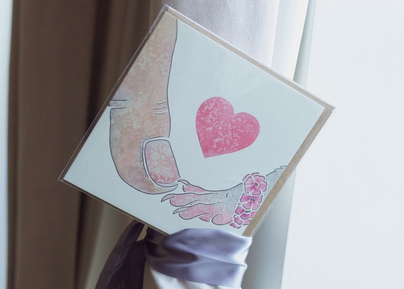 Puede incluir: Una tarjeta de felicitaci&oacute;n cuadrada con una ilustraci&oacute;n en acuarela de un pulgar grande tocando una mano m&aacute;s peque&ntilde;a con garras. Un coraz&oacute;n rosa texturizado se sit&uacute;a entre ambas manos, y la mano peque&ntilde;a lleva una pulsera de flores rosas.