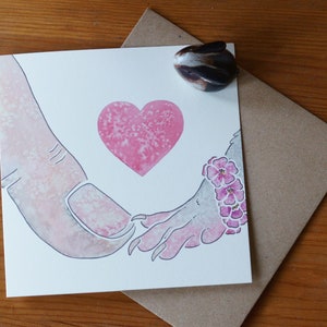 Puede incluir: Una tarjeta de felicitaci&oacute;n cuadrada con una ilustraci&oacute;n en acuarela de un dedo humano grande tocando la mano de una criatura peque&ntilde;a con garras. Un coraz&oacute;n rosa est&aacute; centrado sobre las manos y la criatura lleva una pulsera floral rosa.