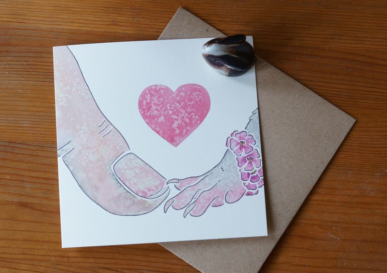 Puede incluir: Una tarjeta de felicitaci&oacute;n cuadrada con una ilustraci&oacute;n en acuarela de un dedo humano grande tocando la mano de una criatura peque&ntilde;a con garras. Un coraz&oacute;n rosa est&aacute; centrado sobre las manos y la criatura lleva una pulsera floral rosa.
