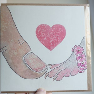 Puede incluir: Una tarjeta de felicitaci&oacute;n cuadrada con una ilustraci&oacute;n en acuarela de un dedo humano grande tocando la mano de una criatura peque&ntilde;a con garras. Un coraz&oacute;n rosa est&aacute; centrado sobre las manos y la criatura lleva una pulsera floral rosa.