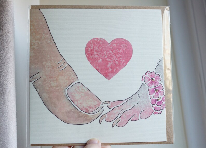 Puede incluir: Una tarjeta de felicitaci&oacute;n cuadrada con una ilustraci&oacute;n en acuarela de un dedo humano grande tocando la mano de una criatura peque&ntilde;a con garras. Un coraz&oacute;n rosa est&aacute; centrado sobre las manos y la criatura lleva una pulsera floral rosa.