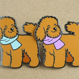May include: Two brown poodle dog enamel pins with a winking face. One pin has a light blue scarf and the other has a light purple scarf.