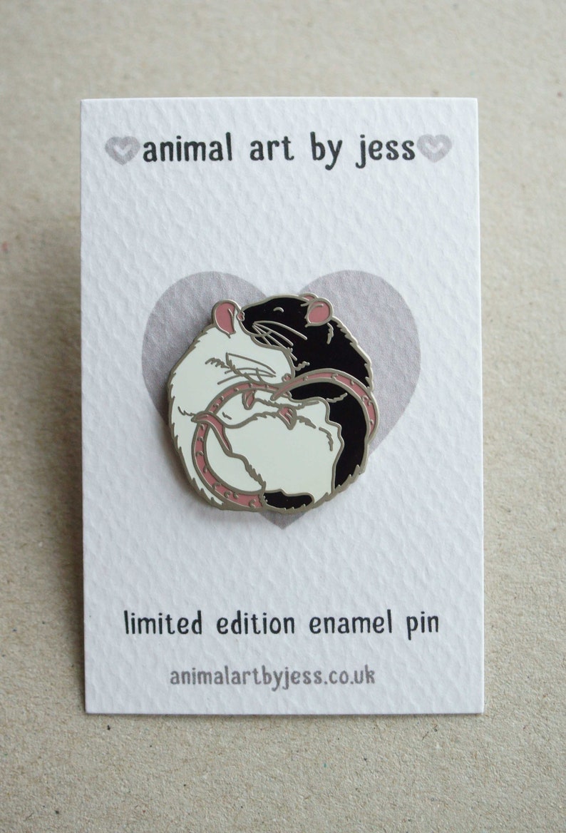An emamel pin attached to a white and grey backing card the size of a business card. The pin shown has one white rat and one black and white hooded rat, with satin nickel plating. The card has a grey heart in the middle where the pin is attached.