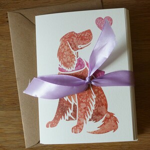 May include: A set of blank greeting cards featuring a watercolor illustration of a dog holding a heart. The cards are tied together with a lavender ribbon.
