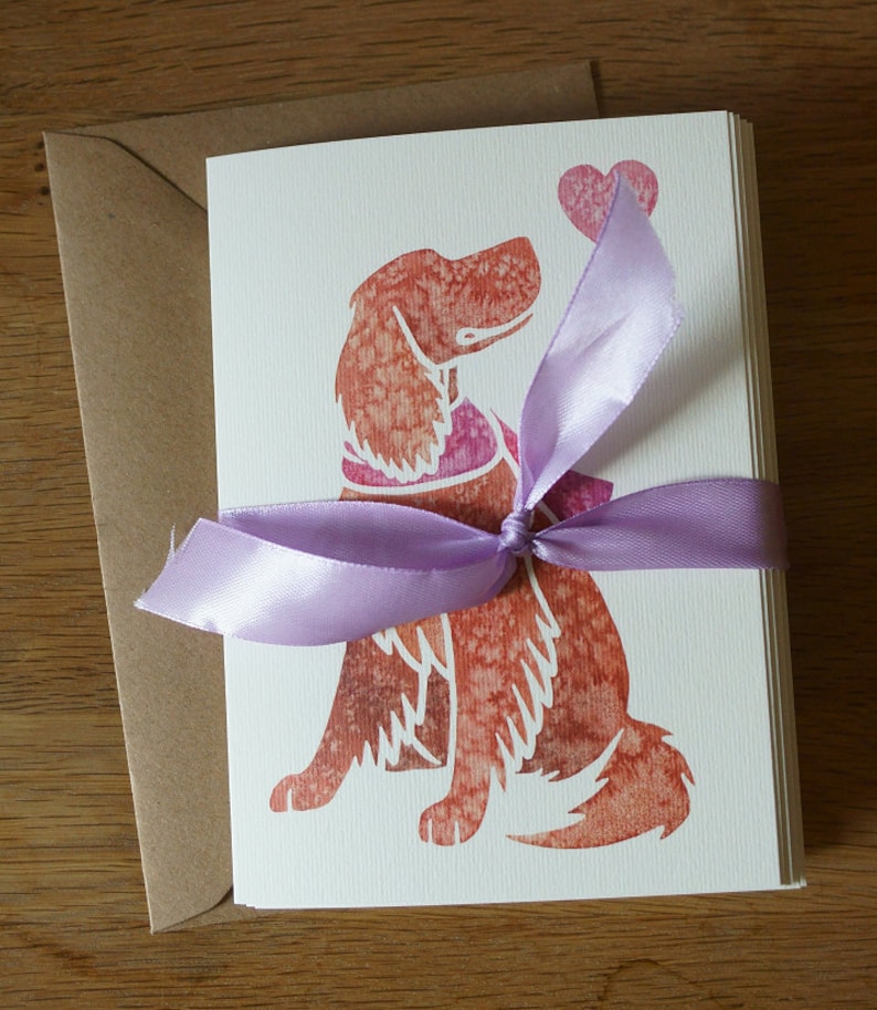 May include: A set of blank greeting cards featuring a watercolor illustration of a dog holding a heart. The cards are tied together with a lavender ribbon.