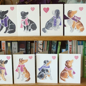 May include: A set of ten greeting cards featuring watercolor illustrations of dogs wearing purple scarves and holding pink hearts. The cards are arranged on a bookshelf.