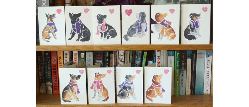 May include: A set of ten greeting cards featuring watercolor illustrations of dogs wearing purple scarves and holding pink hearts. The cards are arranged on a bookshelf.